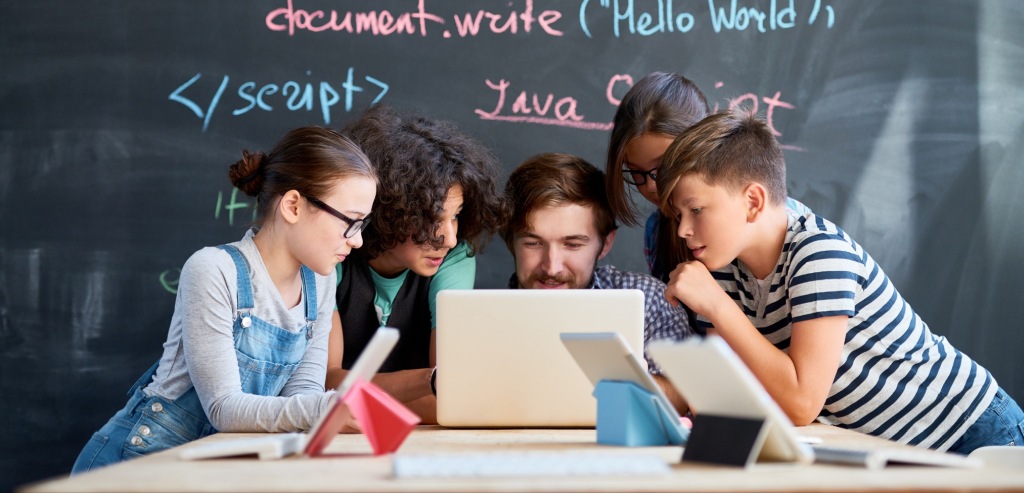 Imagen de portada de sección "Programas" - Se visualizan a jóvenes alrededor de un adulto en un salón de clases, observando una notebook